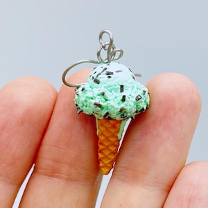 May include: A mint green ice cream cone earring featuring a textured waffle cone base and a scoop of ice cream topped with dark brown chocolate chip sprinkles. The charm is attached to a silver-toned metal earring hook for hanging.