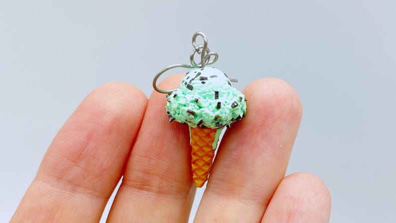May include: A mint green ice cream cone earring featuring a textured waffle cone base and a scoop of ice cream topped with dark brown chocolate chip sprinkles. The charm is attached to a silver-toned metal earring hook for hanging.