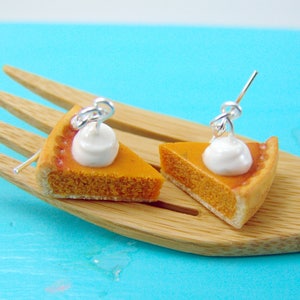 May include: A pair of pumpkin pie earrings with a white whipped cream topping. The earrings are made of polymer clay and are on a silver wire.