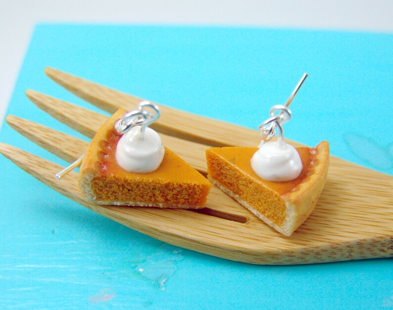 May include: A pair of pumpkin pie earrings with a white whipped cream topping. The earrings are made of polymer clay and are on a silver wire.