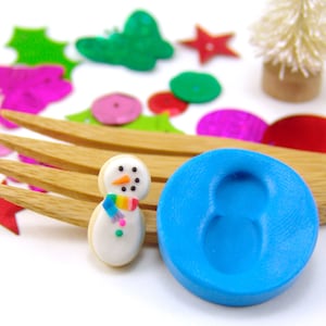 May include: A blue silicone mold shaped like a snowman, next to a decorated snowman cookie. The cookie is white with a rainbow scarf, black eyes, and an orange nose. The mold and cookie are on a wooden surface with colorful confetti and a small white Christmas tree in the background.