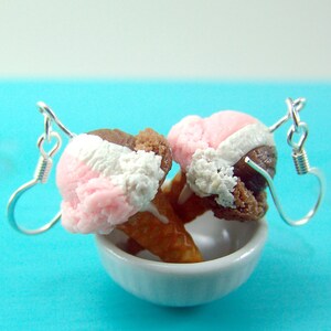 May include: A pair of earrings featuring miniature ice cream cones with pink, white, and brown ice cream. The cones are attached to silver earring hooks.