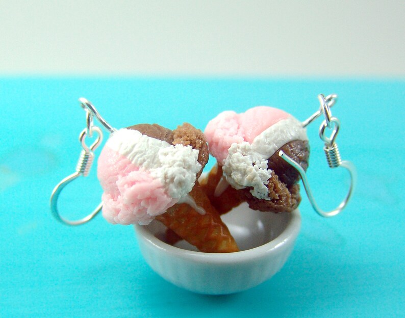 May include: A pair of earrings featuring miniature ice cream cones with pink, white, and brown ice cream. The cones are attached to silver earring hooks.