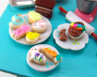 Peut inclure: Un ensemble de biscuits de Pâques miniatures sur des assiettes blanches. Les biscuits sont décorés de couleurs pastel et de motifs variés, notamment des lapins, des poussins et un panier d'œufs.
