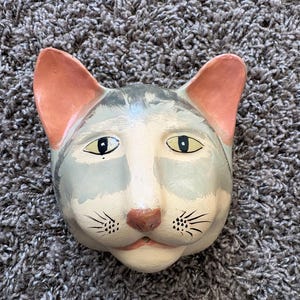 May include: A hand-painted cat face sculpture with a whimsical design. The cat's face is primarily white, gray, and light blue, with pink ears and a brown nose. Black whiskers and yellow eyes complete the design.