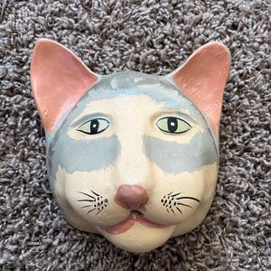 May include: A hand-painted ceramic cat face with a light gray and white color scheme. The cat has pink ears, green eyes, and black whiskers. The cat's nose is a muted pink, and the mouth is a light pink.
