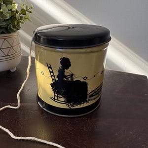 May include: A vintage metal yarn container with a black lid and a yellow body. The container features a black silhouette of a woman knitting on a rocking chair. A white string is attached to the lid. The container is on a dark brown surface.