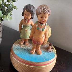May include: A vintage figurine featuring two children in swimwear, adorned with necklaces, standing on a beach-themed base. The base is a wooden-look design with blue and red accents. A small dog figurine is also present.