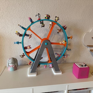 May include: A toy Ferris wheel in orange and teal, with small animal figurines in each carriage. The wheel is supported by grey legs. A pink audio player and a bunny figurine sit on a white surface. The name "Emma" is written on the base.