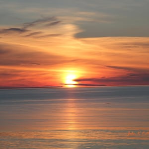 Può includere: Un tramonto panoramico su un corpo d'acqua calmo. Il cielo è pieno di colori vibranti, tra cui sfumature di arancione, rosso e giallo, con alcune nuvole. L'acqua riflette i colori del cielo, creando un paesaggio bello e sereno.