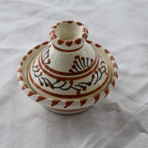 Puede incluir: Una tagine de cerámica blanca con diseños pintados en marrón y negro. La tagine tiene una base redonda y una tapa abovedada.