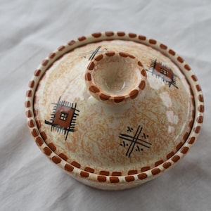 Puede incluir: Una tapa de cerámica blanca con un borde marrón y diseños geométricos. La tapa tiene un pomo en la parte superior.