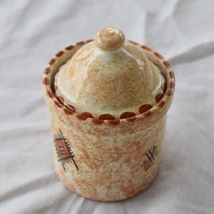 Puede incluir: Un pequeño tarro redondo de cerámica con tapa. El tarro es de color beige con motas marrones y detalles decorativos marrones. La tapa tiene un pequeño pomo. El tarro tiene un diseño geométrico en el lateral.