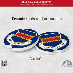 May include: Two round ceramic sandstone car coasters featuring a blue shield with a red zigzag stripe and the Latin text "VIDEMUS OMNIA" on a white scroll. The coasters have a small notch on the edge and are white on the sides and bottom.