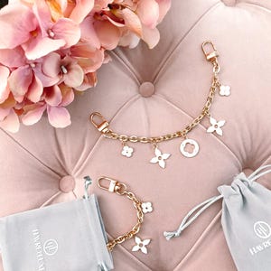 May include: Two gold chains with white flower charms and a Louis Vuitton logo charm. The chains are on a pink velvet surface with two grey velvet bags with the text "HAVREFLUXE" on them.