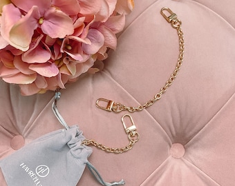 May include: Two gold chains with clasps, one longer than the other, on a pink velvet surface. A small gray velvet bag with the text "HAVREFILLUXE" and a logo is also on the surface.