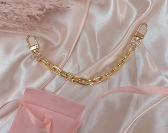 May include: Gold-colored chain with lobster claw clasps, displayed with a pink satin pouch and ribbon. The chain is made of rectangular links. The pouch has the text "HAVRELINE" printed on it. The background is a pink satin fabric.