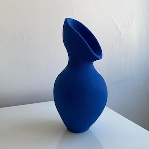 Puede incluir: Un jarrón de cerámica azul cobalto con un diseño asimétrico único. El jarrón tiene una base redondeada que se estrecha hacia un cuello delgado, que luego se ensancha en una abertura ancha y angular. La superficie tiene un acabado mate.