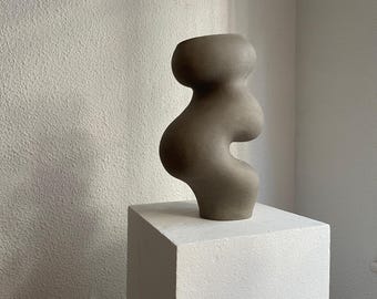 May include: A gray ceramic vase with an abstract, organic shape. The vase is sitting on a white pedestal.