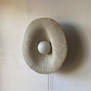 May include: A textured wall sconce featuring a central white globe light. The sconce has an organic, oval shape and a neutral, off-white colour. A small chain hangs from the bottom.