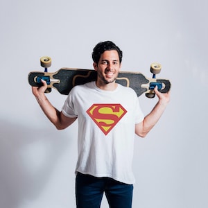 May include: A white t-shirt with a red and yellow Superman logo. The person is holding a skateboard behind their back.
