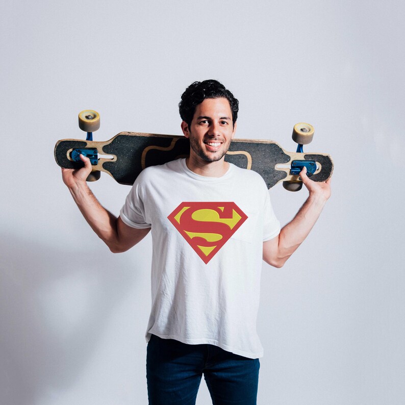May include: A white t-shirt with a red and yellow Superman logo. The person is holding a skateboard behind their back.