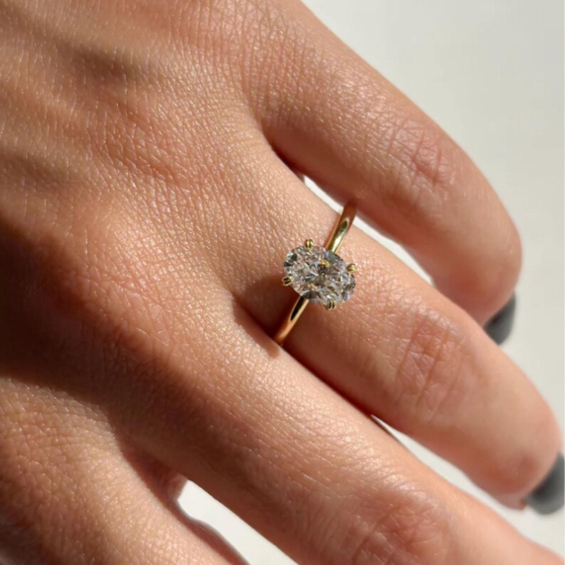 May include: A gold ring featuring a large, oval-cut diamond. The diamond is the focal point, set in a classic four-prong setting. The band is a thin, polished gold. The ring is shown on a person's finger.
