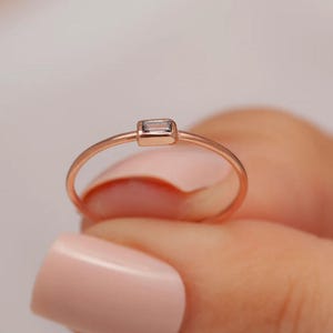 Op de afbeelding: Een roségouden ring met een kleine, rechthoekige, heldere edelsteen. De ring wordt tussen vingers gehouden met lichtroze nagellak.