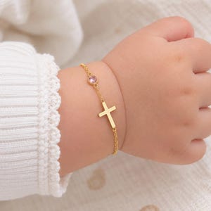 Puede incluir: Una delicada pulsera de cadena en tono dorado que presenta un pequeño dije de cruz centrado y una piedra preciosa redonda de color rosa claro engastada en un bisel dorado. La pulsera tiene una cadena de metal pulido.