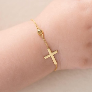 Puede incluir: Un brazalete dorado con un colgante de cruz. El brazalete tiene una cadena delicada con una pequeña piedra redonda. La cruz es de diseño simple y clásico, y el brazalete se muestra en una muñeca.
