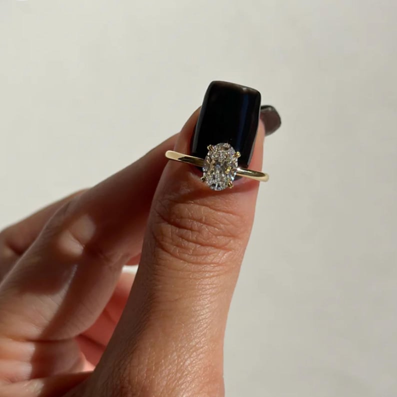 May include: A gold ring featuring an oval-cut diamond. The ring's band is thin and delicate, and the diamond is set in a classic prong setting. The ring is shown against a neutral background.