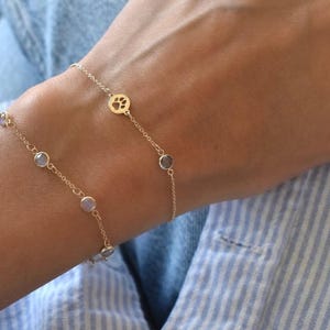 Può includere: Due delicati braccialetti d'oro. Uno presenta un ciondolo a forma di zampa, l'altro ha piccole pietre rotonde di colore chiaro. I braccialetti sono indossati su un polso, su uno sfondo di camicia a righe blu e bianche.