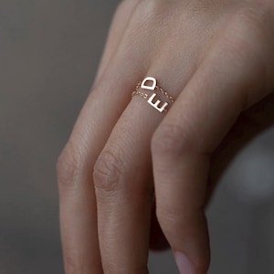 May include: A delicate gold chain ring featuring two small, uppercase metal letters, D and E, suspended from the fine link band. The ring is crafted from a polished gold-tone metal, showcasing a minimalist design with the letters positioned vertically.