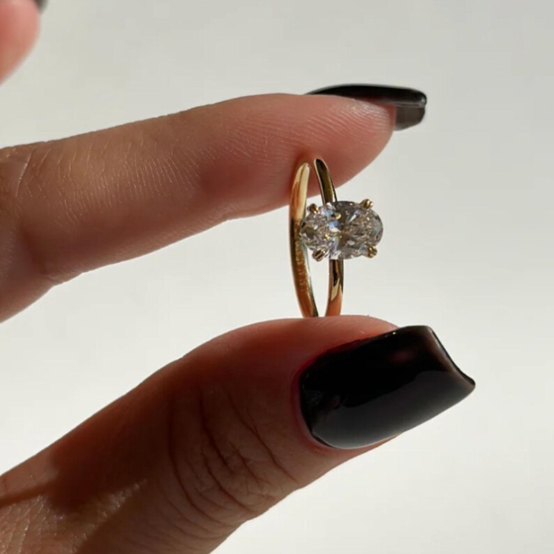 May include: A gold ring with a large, oval-shaped, clear gemstone. The ring is held between two fingers against a white background. The ring's band is thin and polished.