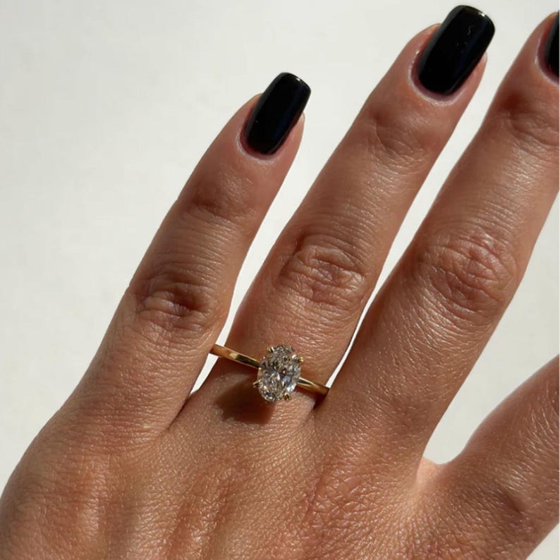 May include: A gold ring with a large oval-cut diamond. The ring is set on a thin gold band. The hand has black nail polish.
