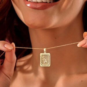 Puede incluir: Collar de cadena dorada con un colgante cuadrado con las letras "SA" en una fuente decorativa.