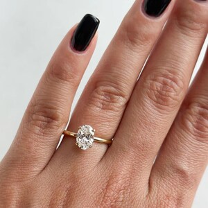 May include: A gold ring with a large, oval-shaped diamond. The diamond is clear and sparkling, set on a thin, yellow gold band. The ring is shown on a hand with black nail polish.
