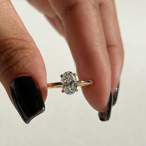 May include: A gold ring with an oval-cut diamond. The diamond is set in a four-prong setting. The ring is held between two fingers with black nail polish.