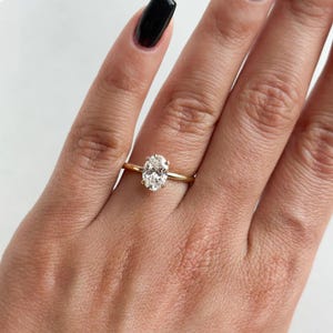 May include: A gold engagement ring with an oval-cut diamond. The ring is set on a thin gold band. The ring is on a person's finger, with the hand and fingers visible.