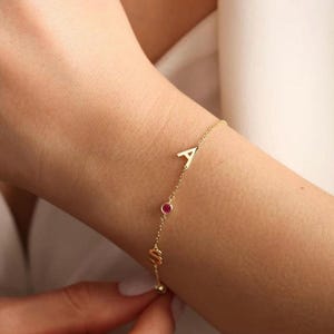 May include: A gold chain bracelet with three charms: a letter A, a letter S, and a small red gemstone.