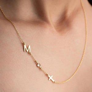 Può includere: Una delicata collana d'oro con la lettera "M", una piccola gemma e un ciondolo a forma di croce. La collana è mostrata su uno sfondo color carne, evidenziando i dettagli raffinati e l'elegante design del gioiello.