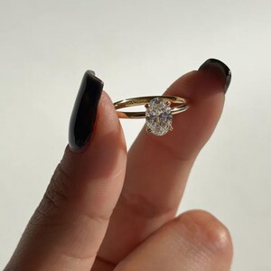 May include: A gold engagement ring with an oval-cut diamond. The ring has a thin band and a four-prong setting. The diamond is clear and sparkling, set against a white background.