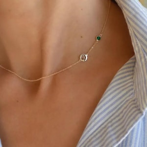 May include: A delicate gold necklace featuring a paw print charm and a small green gemstone. The necklace is worn on a person, with a striped blue and white shirt in the background. The necklace is a subtle and elegant piece of jewelry.