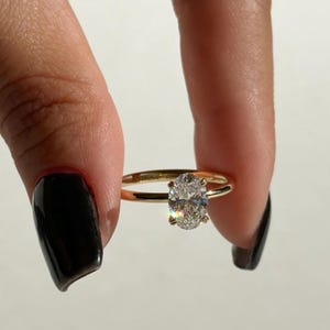 May include: A gold ring with a large, oval-cut diamond. The ring is held between two fingers against a white background. The diamond sparkles, reflecting light. The band is thin and simple.