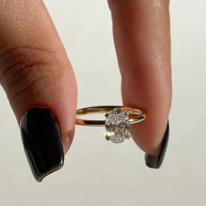 May include: A gold ring with a large, oval-cut diamond. The ring is held between two fingers against a white background. The diamond sparkles, reflecting light. The band is thin and simple.