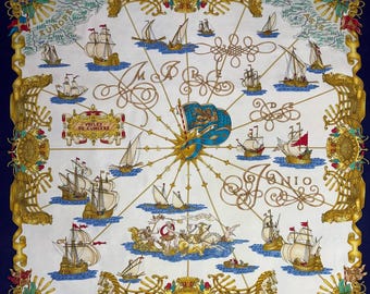 May include: A square silk scarf with a navy blue border and a white centre featuring a nautical theme. The design includes sailing ships, a compass rose, and the text "VOILES DE LUMIERE" in gold lettering. Decorative gold filigree frames the design.