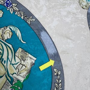 May include: A silk scarf with a teal and grey border, adorned with floral and classical figure illustrations. The scarf has a cream-coloured, textured background. Blue roses and green leaves accent the design.