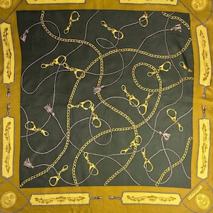 May include: Square silk scarf with a dark green center and a gold border. The scarf features a repeating pattern of gold chains, clasps, and tassels. The border has decorative rectangular panels and a circular emblem.