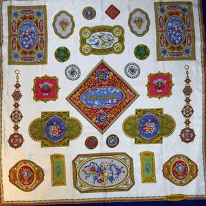 May include: A white silk scarf with a repeating pattern of floral and ornamental designs in red, blue, green, and gold. The scarf features the brand name "Trussardi" and decorative borders.