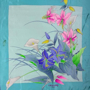 May include: A square silk scarf with a floral design. The scarf features pink lilies, purple irises, and white calla lilies against a light blue background. The border is a darker shade of blue. The brand name "LEONARD PARIS" is printed in the bottom right corner.